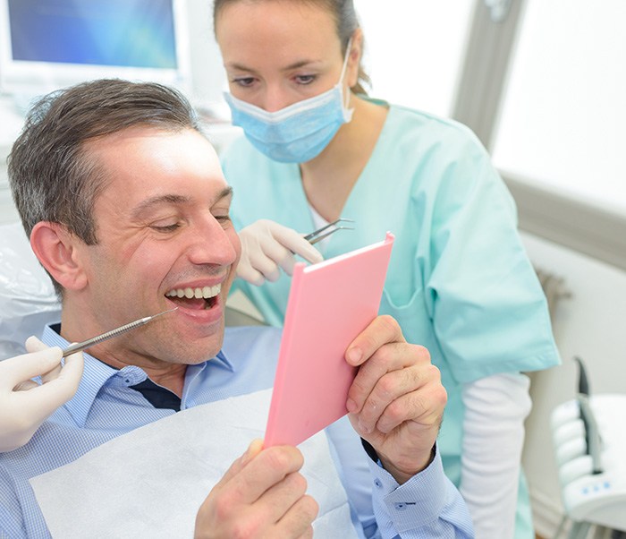 Man in dental chair smiling at results in handheld mirror with dentist looking over his shoulder