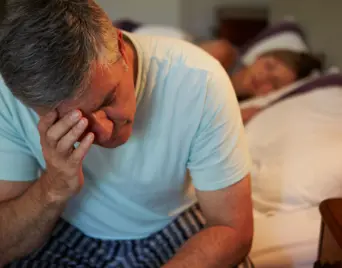 Man sitting awake in bed and rubbing his eyes next to his sleeping partner
